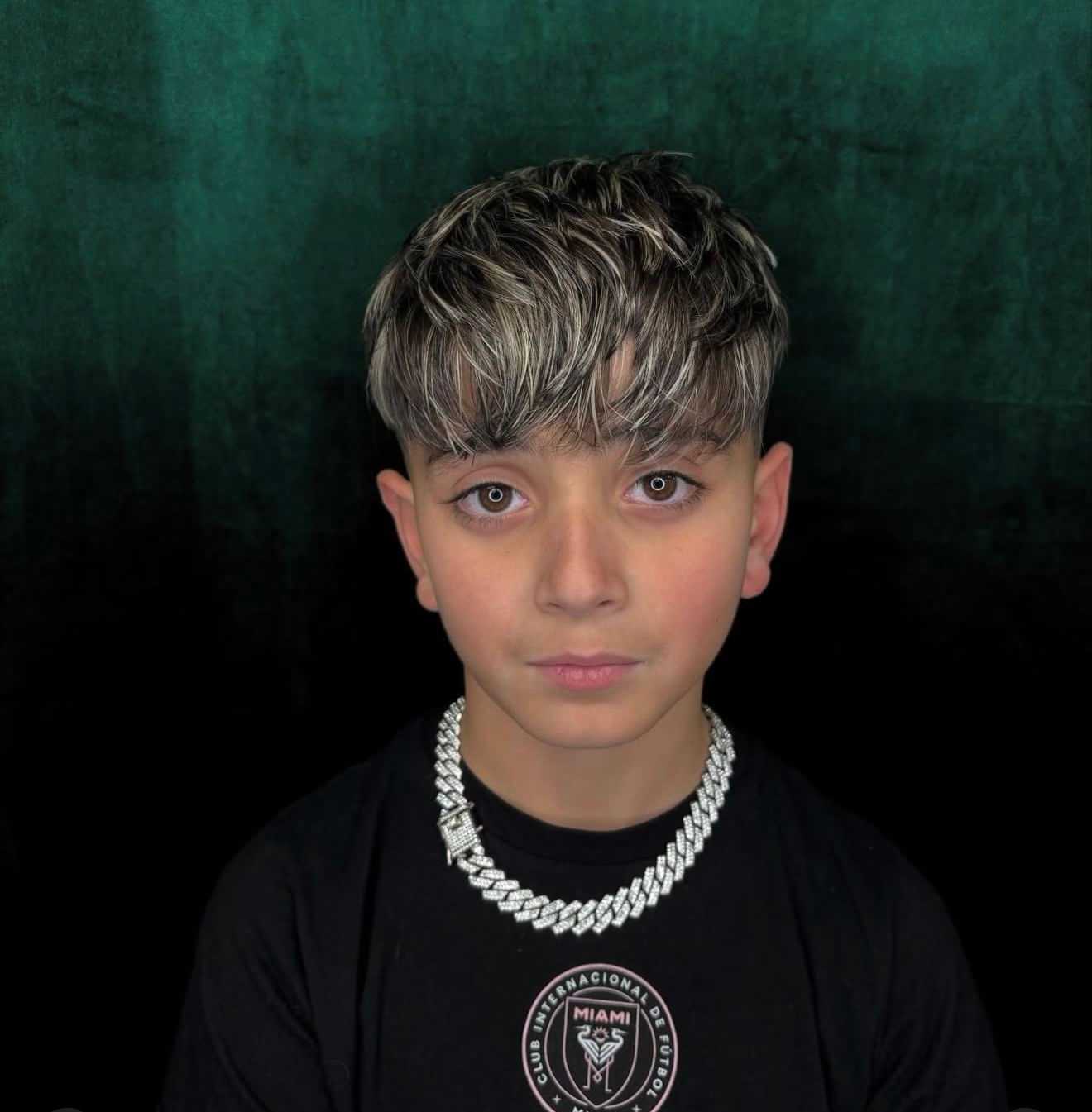 Boy with stylish haircut and silver chain, wearing a black T-shirt against a dark background.