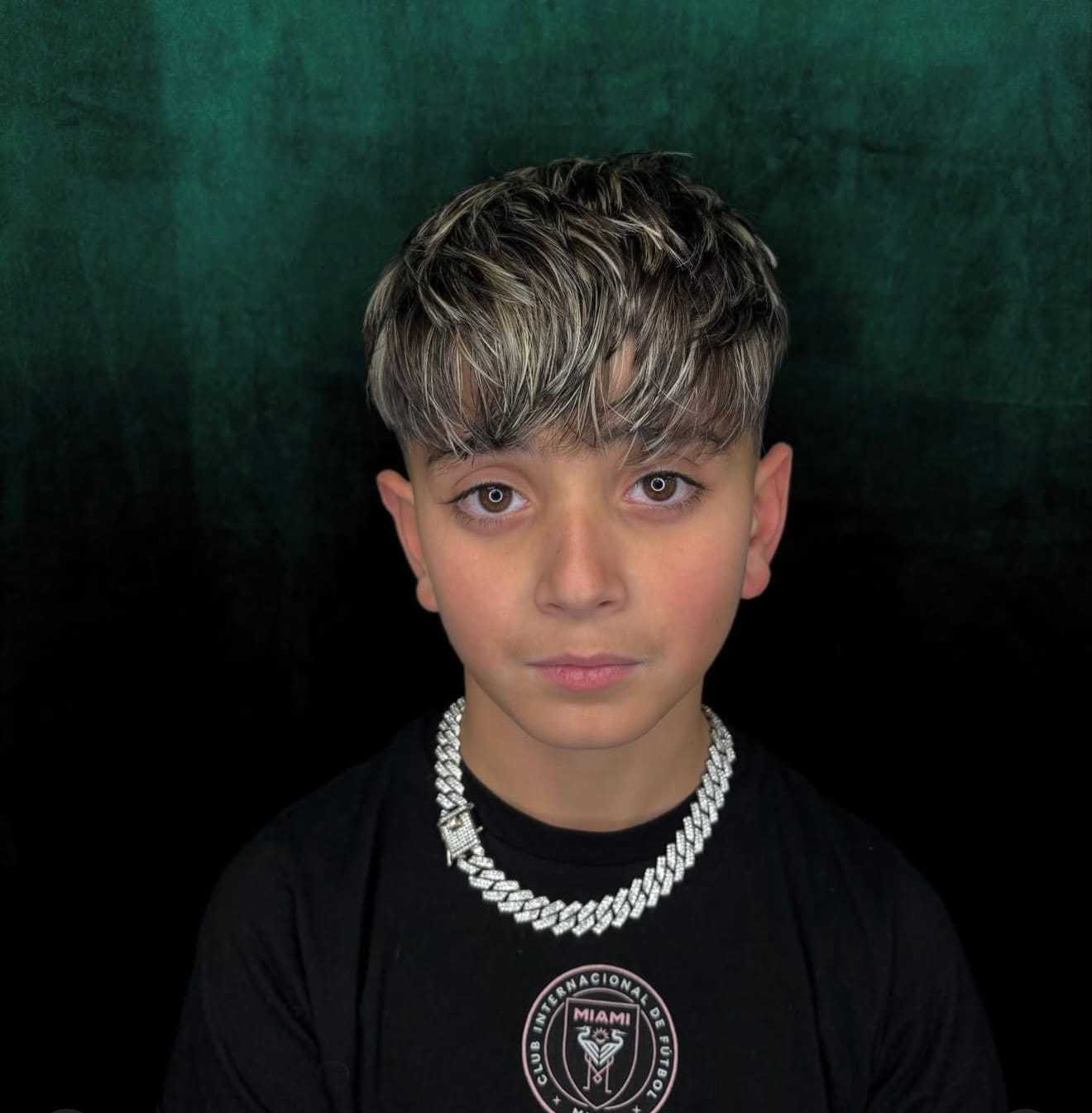 Boy with stylish haircut and silver chain, wearing a black T-shirt against a dark background.