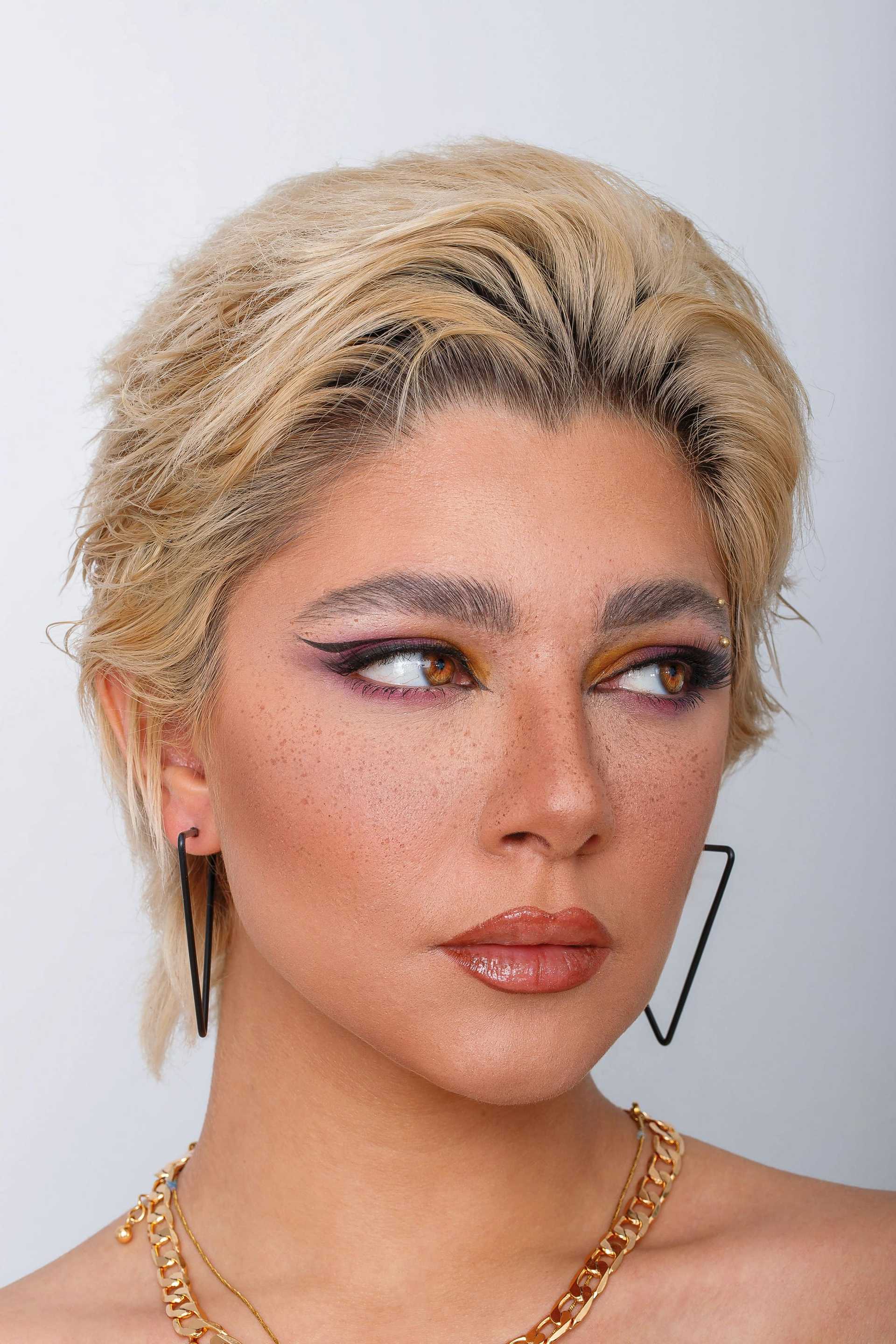 Person with short blond hair, bold makeup, hoop earrings, and gold necklace against gray background.
