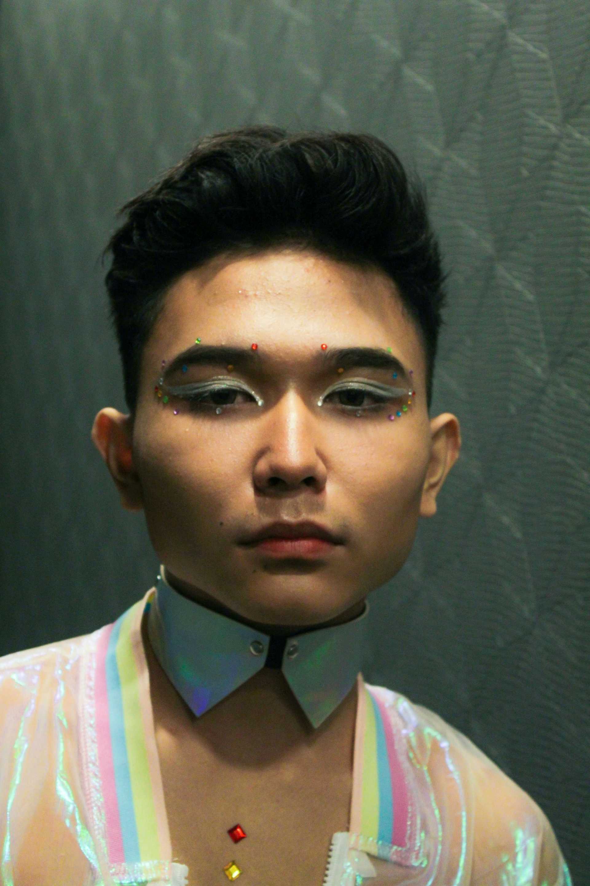 Young person with colorful makeup and holographic outfit in a reflective setting.